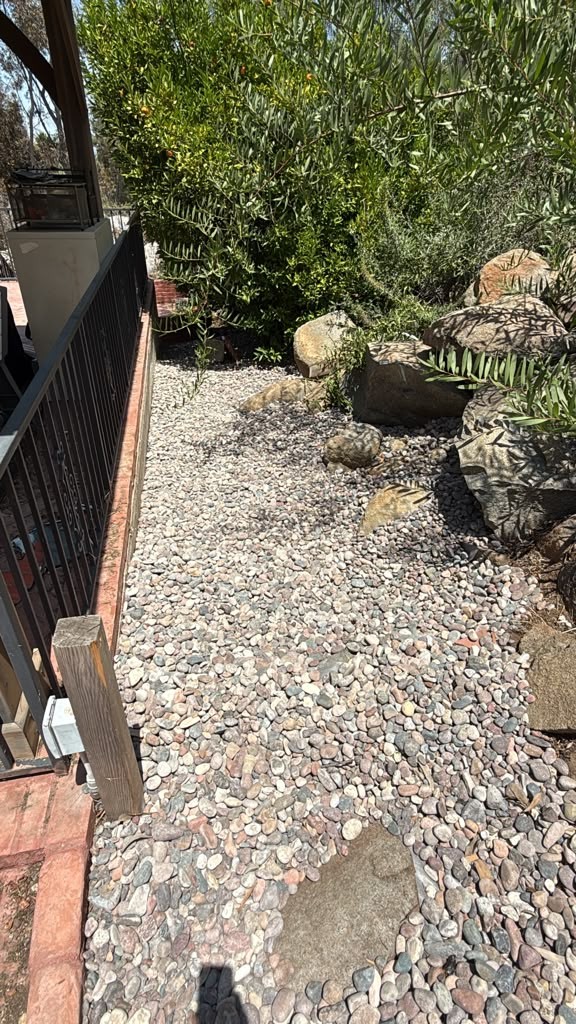 A river-rock path beside a railing and brick edging, with boulders and shrubs along one side, in the El Cajon yard