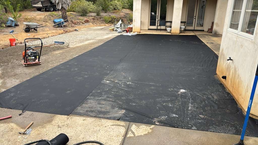 Black weed-barrier fabric laid over the compacted base beside the house, with a plate compactor on the gravel